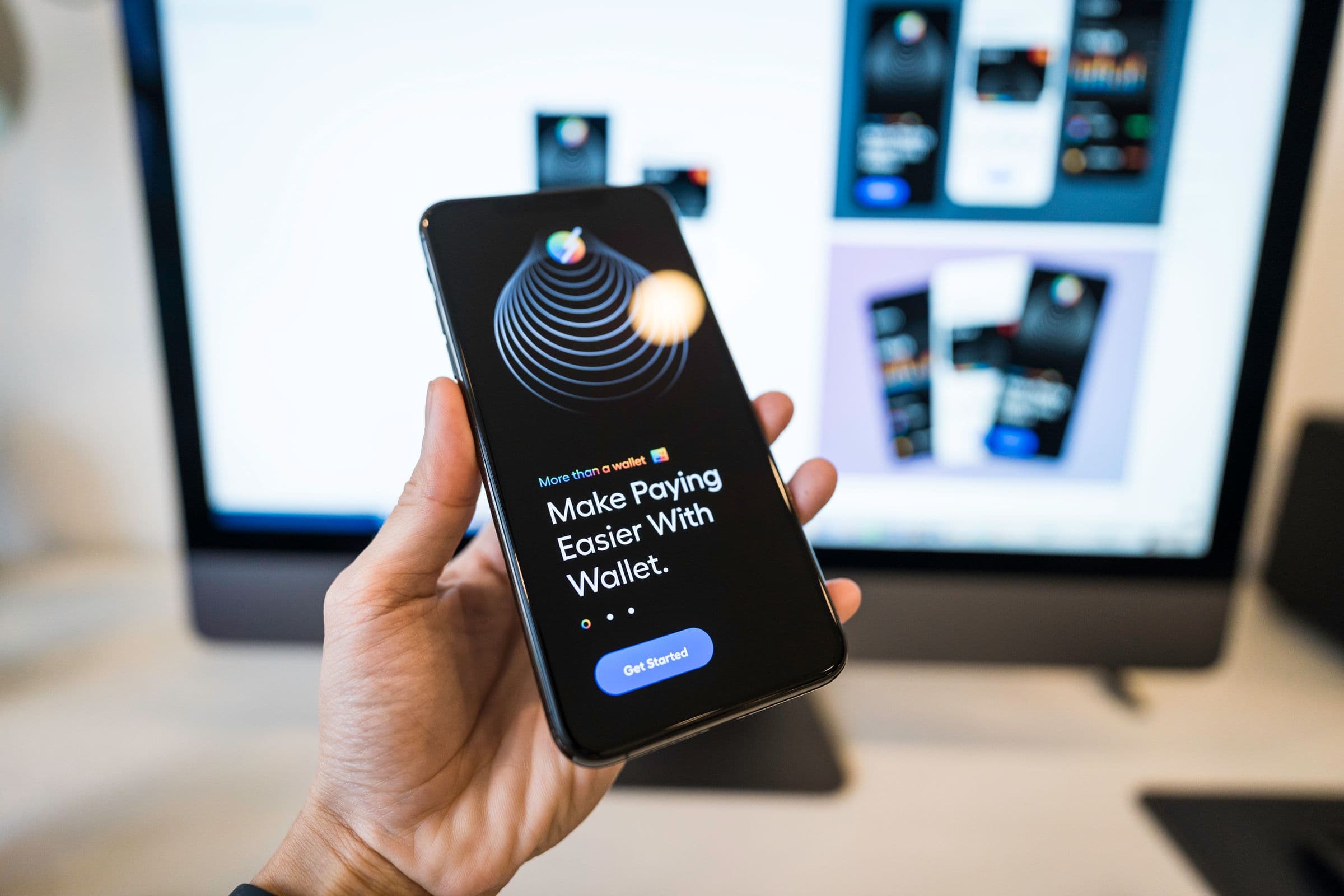 A hand holding a phone with an app open; the app’s design screens on the monitor behind